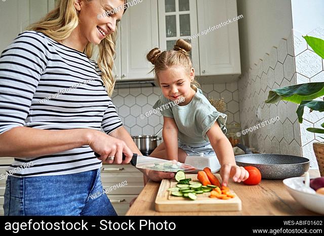 Smiling mother cutting vegetables by daughter sitting on kitchen counter