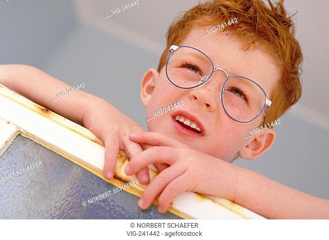 portrait, close-up, funny redheaded boy with freckles and big spectacles with blue frame, 6 years, wearing white shirt and blue trousers with flap looking...