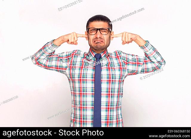 I don't want to hear. closed eyes unhappy bearded businessman in colorful checkered shirt, blue tie and eyeglasses standing with finger in ears