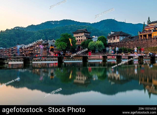 Feng Huang, China - August 2019 : People walking on the narrow wooden passage across Tuo river, flowing through the centre of Feng huang Old Town on a calm...