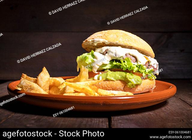 Delicious gourmet beef burger with creamy sauce, lettuce, tomato and guacamole, served with french fries on a rustic plate on a wooden table