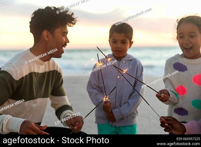 Multiracial children holding sparklers enjoying with parents at beach against sky during sunset
