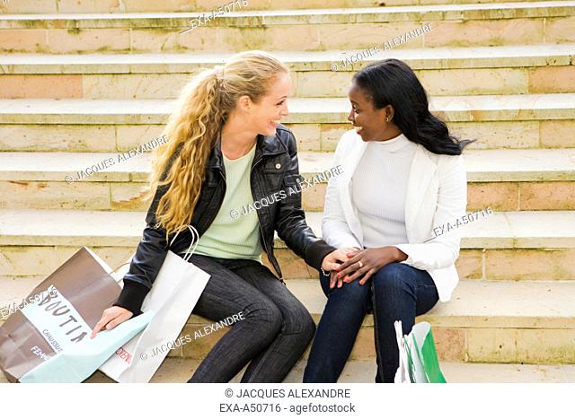 Caucasian and african woman sitting on stairs in the city after having completed their shopping