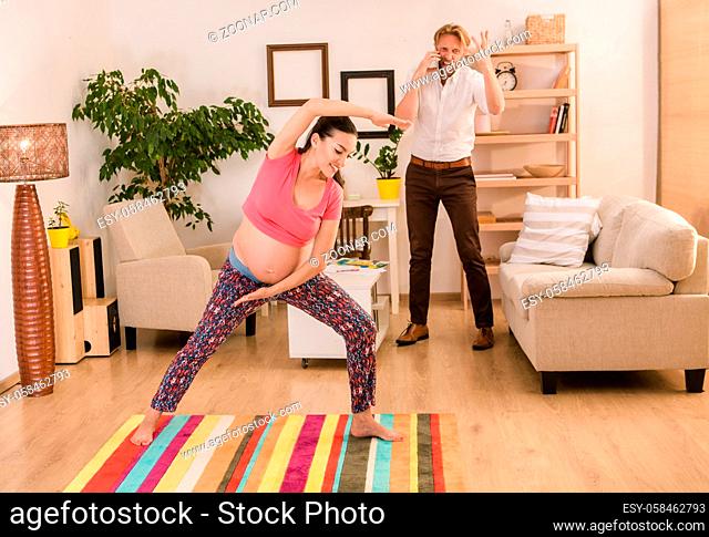 Calm and relaxed pregnant woman having yoga exercises at home while her husband screaming and shouting solving some business issues or problems on background