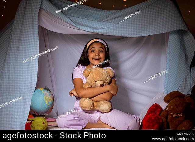 Little girl embracing teddy bear while sitting inside a tent in bedroom