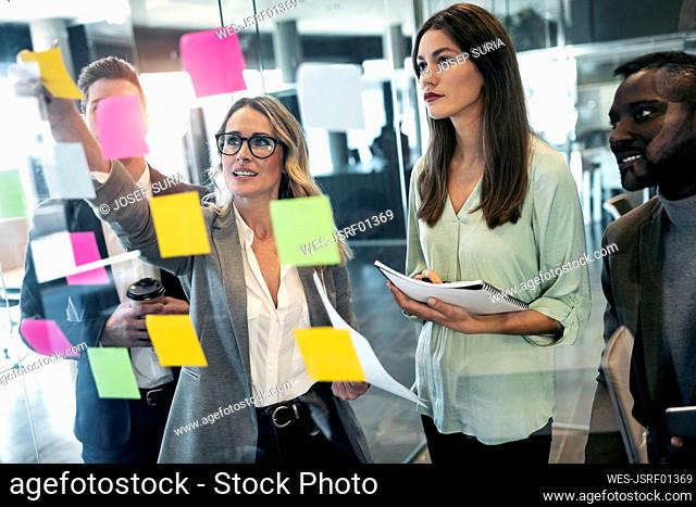 Businesswoman discussing over adhesive note on glass wall while standing with colleague in office