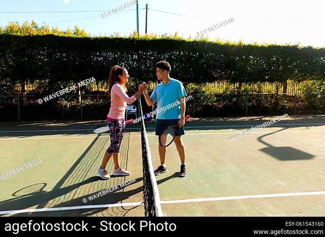 Smiling biracial couple playing tennis shaking hands over the net on sunny outdoor tennis court