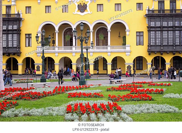 Plaza del Armas: detail of City Hall, yellow stucco and wooden balconies typical of Spanish Colonial era, landscaped gardens with red flowers, Lima, Peru