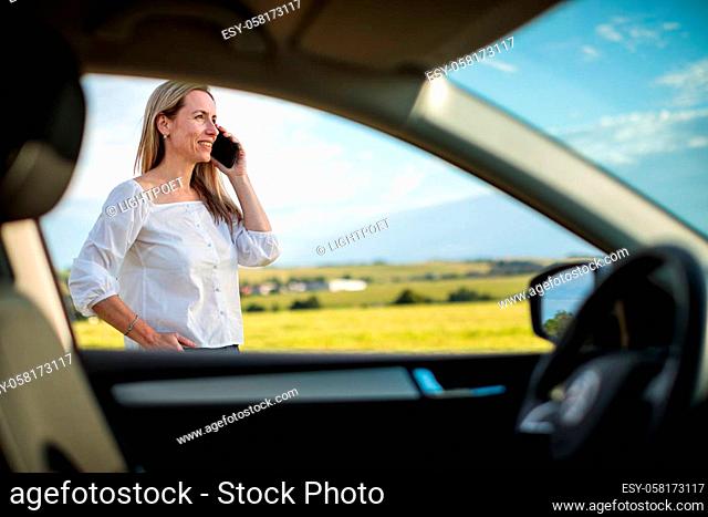 Pretty midle aged woman at the steering wheel of her car, having a break on a long trip, making a phone call