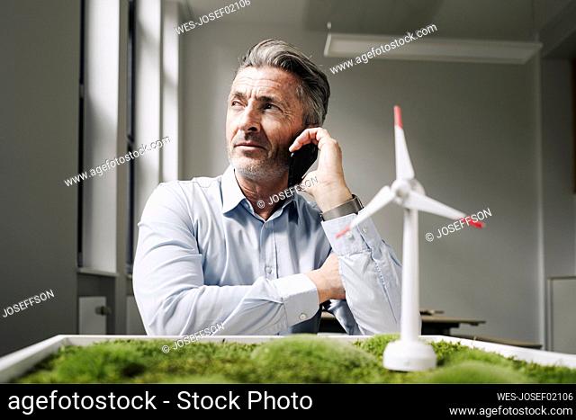Businessman on the mobile phone sitting by moss frame and wind turbine toy at office