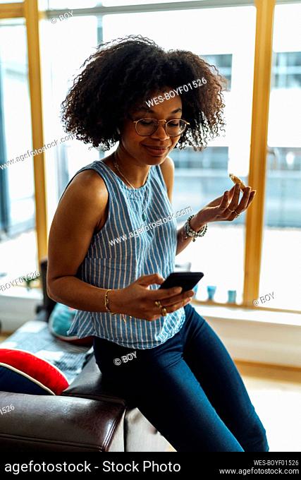 Female entrepreneur holding bread using smart phone while sitting on sofa at home