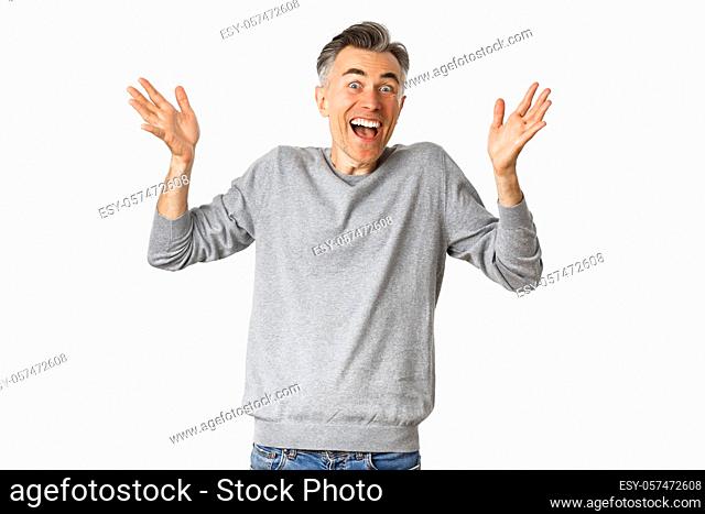 Image of excited and happy middle-aged man with gray short hairstyle, raising hands up and cheering, standing over white background