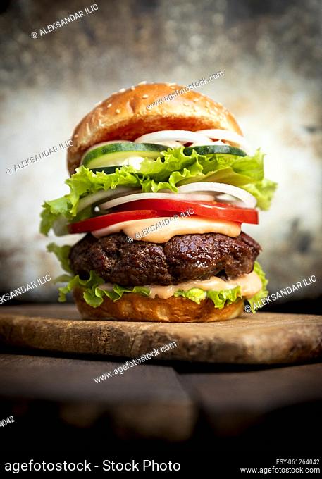 Burger with beef meat, cheese, lettuce, onion, tomato, cucumber, mayo and ketchup on an old wooden table. Junk and unhealthy food concept