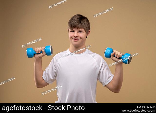 Waist-up portrait of a pleased adolescent boy holding a pair of dumbbells with arms bent