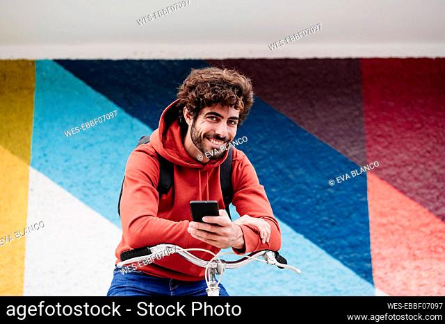 Smiling man with smart phone on bicycle in front of colorful wall