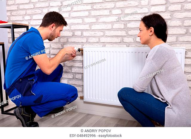 Beautiful Woman Looking At Young Male Plumber In Uniform Installing Radiator With Screwdriver