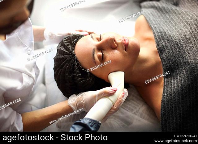 Female beautician in mask doing laser therapy procedure on face of relaxed woman during work in cosmetology clinic