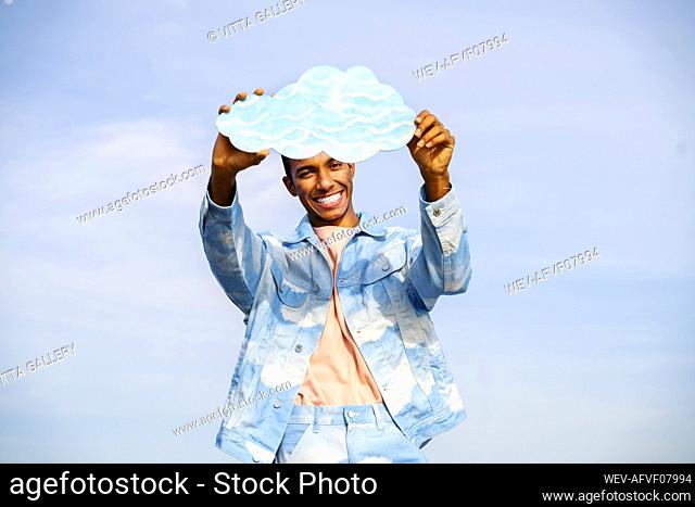 Smiling young man holding cloud while standing against sky