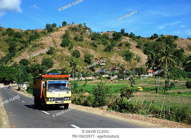 Truck leaving village on highway with small terraced ricefields and other crops sits below hills with bushes and scrub Sape Sumbawa Timur NTB Indonesia