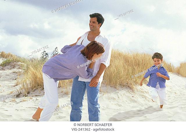beach-scene, full-figure, dark-haired family with 4 year old son is playing bare feeted in the dunes, woman mid of 20 wearing white trouser and blue shirt and...