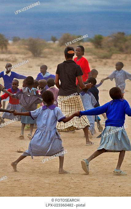 Masai school out on the plains: a group of children, mostly dressed in blue sweaters, and blue check clothing dance in a circle around their two teachers