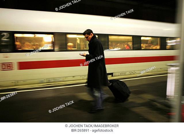 DEU Germany : Man walks with a mobile phone and a suitease trolley on a platform of a railway station. |