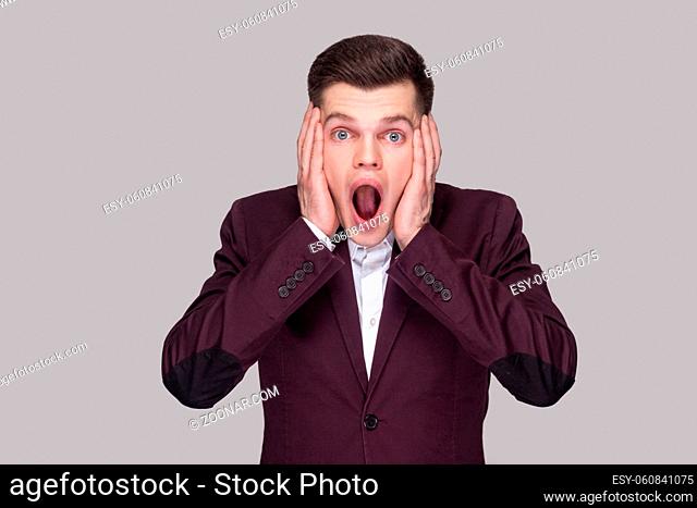 Portrait of amazed handsome young man in violet suit and white shirt, standing, looking at camera and holding hands on face with shocked surprised face
