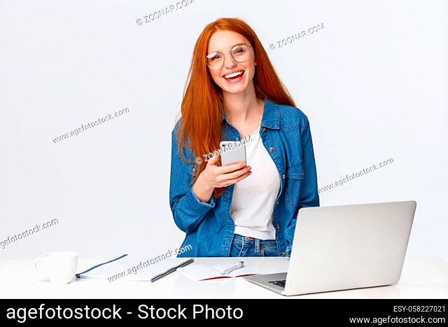 Cheerful, friendly-looking charismatic redhead female designer, freelancer with red foxy hair, glasses, laughing carefree, holding smartphone