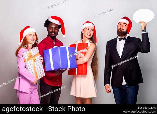 Well dressed man looking away with puzzlement and dreaming face, her friends standing near and holding gift box. Studio shot. Gray background