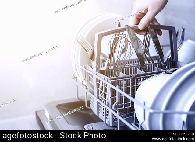 Close-up of female hand holding full cutlery basket with clean knife, fork, whisk. Loading to, empty out or unloading from open automatic dishwasher machine...