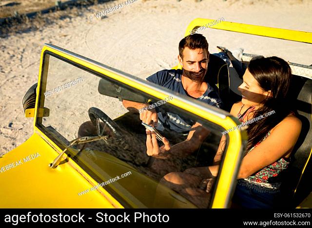 Happy caucasian couple sitting in beach buggy by the sea using smartphone