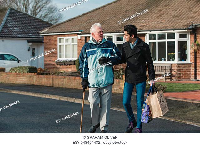 Teenage boy is walking back from the shop with his grandparent. He is carrying the shopping bag and they are linking arms