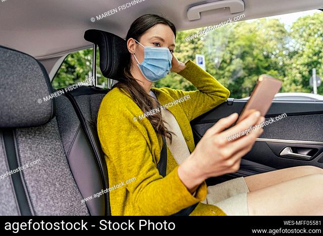Young woman wearing protective mask sitting on back seat of car looking at cell phone