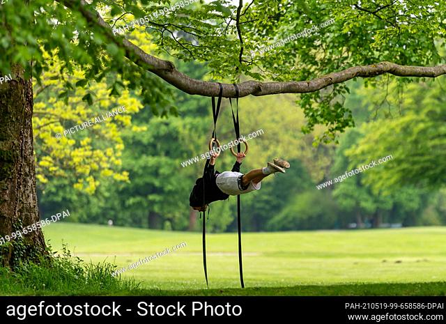 19 May 2021, Bavaria, Munich: Calisthenics trainer Alexander Wujkov hangs with his gymnastic rings from a long tree branch in the English Garden