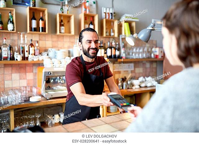 small business, people and service concept - happy man or waiter in apron with card reader and customer paying at bar of coffee shop