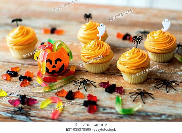 food, baking and holidays concept - cupcakes or frosted muffins with halloween party decorations and candies on wooden table