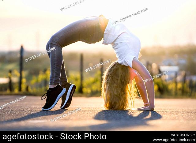 Young flexible woman doing backward bend on road during sunny day