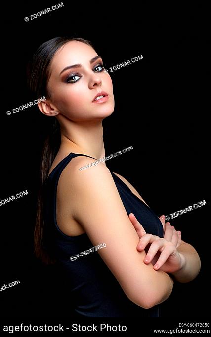 Beauty portrait of young woman in black dress with hands crossed. Brunette girl with evening female makeup on black background