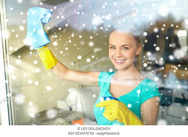 people, housework and housekeeping concept - happy woman in gloves cleaning window with rag and cleanser spray at home over snow effect