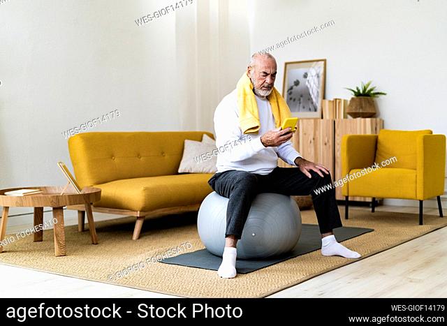 Senior man using mobile phone on fitness ball in living room