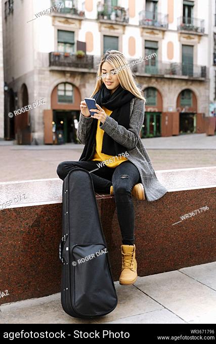 Young woman with violin case using smartphone in city