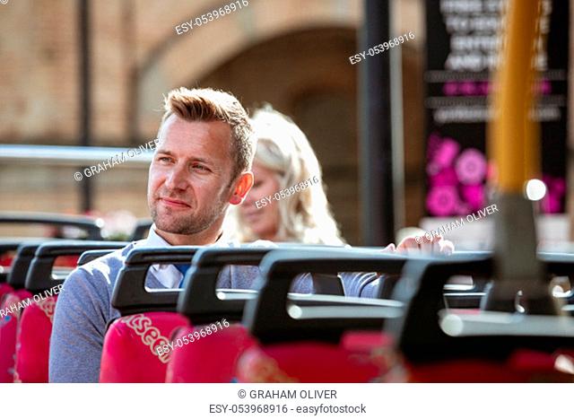 A businessman sitting by himself on an open-air bus admiring the view. There is an unrecognisable woman behind him