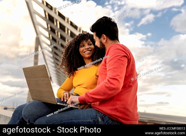 Happy young couple sitting with laptop under cloudy sky