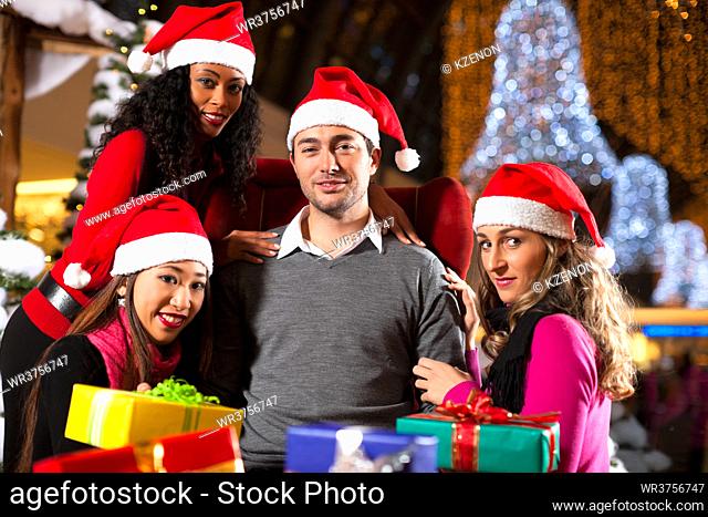 Group of four people - diversity - with Santa hats sitting amid artificial snow covered fir trees and lights with Christmas presents in a shopping mall