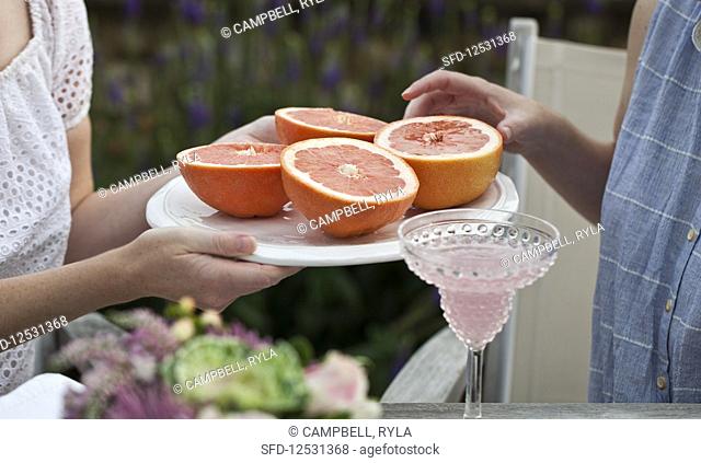 Two women sitting at an outdoor brunch, one woman passing a tray of grapefruit halves and the other woman reaching for one