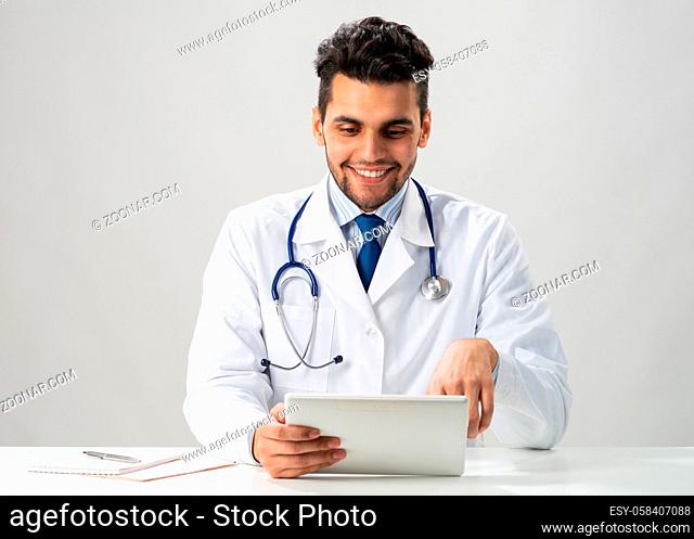 Young smiling internist using tablet computer. Serious therapist in white medical gown with stethoscope sitting at desk. Modern digital technology in diagnosis...
