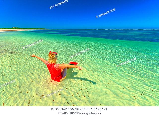 Freedom woman with snorkeling wetsuit, mask and fins pink and peach color, sitting on the seashore with oper arms. Female looking tropical sea in Hangover Bay