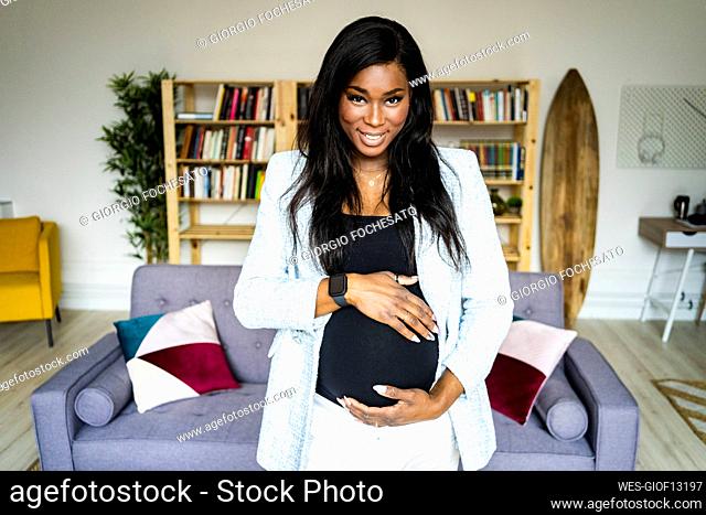 Smiling young pregnant businesswoman touching abdomen in living room at home