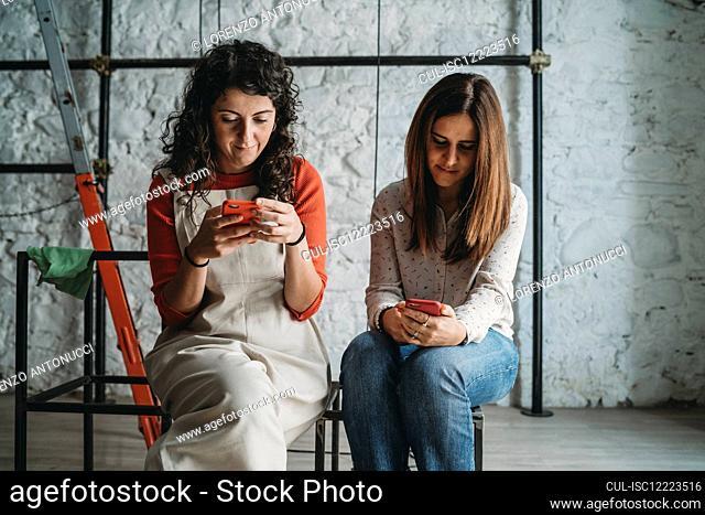Two mid adult women looking at smartphones in their new shop
