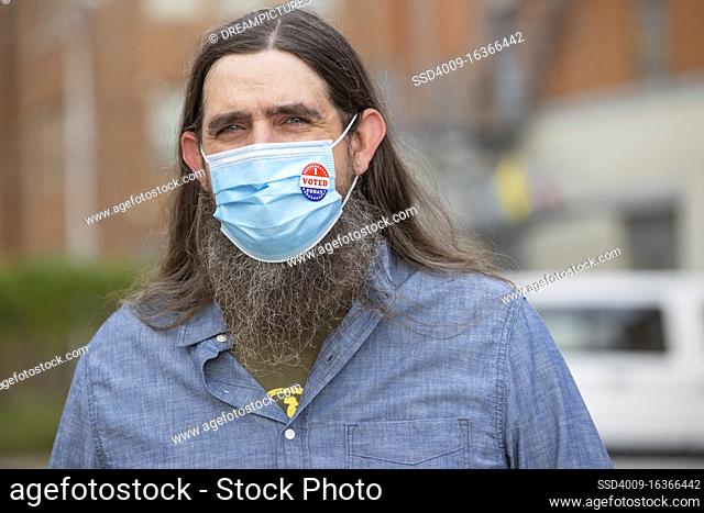 Caucasian male with long hair and a full beard wearing a mask and an I Voted Today sticker on the mask, looking into camera